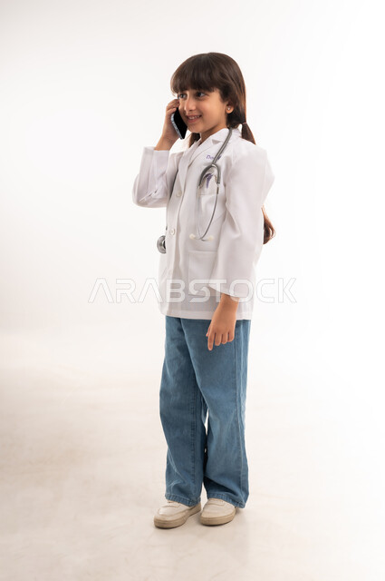 استخدام التكنولوجيا لتقديم الإستشارات الطبية، بورتريه لفتاه عربية خليجية عمانية ترتدي المعطف الطبي والسماعات، بنت سعودية تقوم بإجراء مكالمة هاتفيه عبر الهاتف المحمول، تلبية مطالب الرعاية الصحية للمرضى عن بعد، صورة الجسم بالطول الكامل، خلفية بيضاء