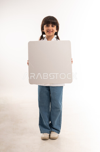 مفهوم العمل في مجال الطب والرعاية الصحية، مهن ووظائف مستقبلية، بورتريه لفتاة عربية خليجية عمانية مبتسمة ترتدي المعطف الطبي وتنظر إلى الكاميرا، بنت سعودية تحمل بيدها لوحة بيضاء فارغة، عرض موك أب مربع أبيض فارغ، الطول الكامل للجسم، خلفية بيضاء