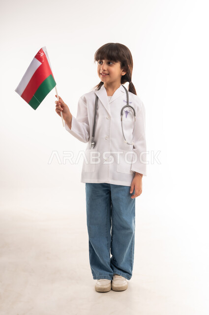 الطموحات والأهداف المستقبلية بالعمل في مستشفيات سلطنة عمان، بورتريه لفتاة عربية خليجية عمانية مبتسمة ترتدي زي الطبيب والسماعات الطبية تحمل بيدها العلم العماني، الفخر والاعتزاز بالانتماء للهوية الوطنية, تحديد المهنة منذ الصغر، الطول الكامل للجسم، خلفية بيضاء