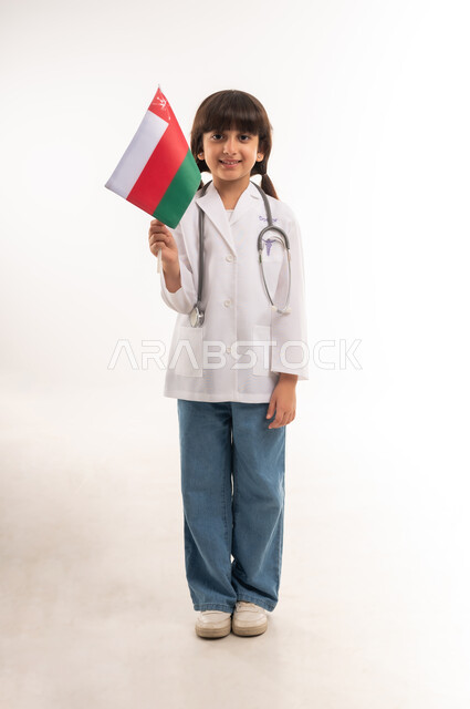 الأهداف والطموحات المستقبلية بالعمل في مستشفيات سلطنة عمان، بورتريه لفتاة عربية خليجية عمانية مبتسمة ترتدي زي الطبيب والسماعات الطبية تحمل بيدها العلم العماني، الفخر والاعتزاز بالانتماء للهوية الوطنية, تحديد المهنة منذ الصغر، الطول الكامل للجسم، خلفية بيضاء