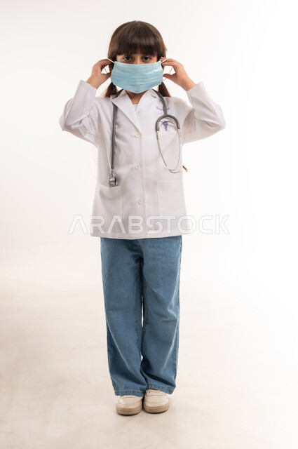 الاحترازات الوقائية ضد كوفيد 19، مفهوم الطب والرعاية الصحية، طموحات وأهداف مستقبلية في العمل في المستشفيات، بورتريه لفتاة عربية خليجية عمانية ترتدي زي الطبيب، بنت سعودية تقوم بوضع الكمامة، اختيار وتحديد مهنة المستقبل منذ الصغر، الطول الكامل للجسم، خلفية بيضاء