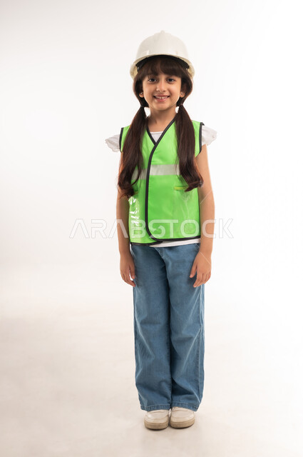 Gestures of passion to achieve childhood dream, work in engineering sector, choosing future since childhood, smiling Saudi girl standing straight, portrait of an Arab Gulf Omani girl wearing jacket and protective helmet for engineers, full body length, white background