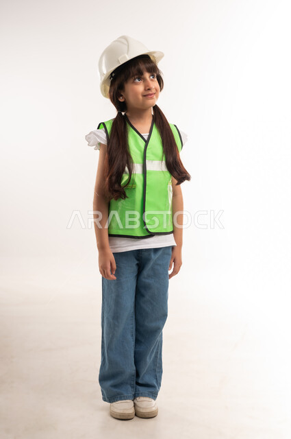 النظر إلى الأعلى بإيماءات الشغف بالوصول إلى حلم الطفولة، العمل في القطاع الهندسي، اختيار المستقبل منذ الصغر، بنت سعودية تقف باستقامة، بورتريه لفتاة عربية خليجية عمانية ترتدي الستره وخوذة الحماية الخاصة بالمهندسين، الطول الكامل للجسم، خلفية بيضاء