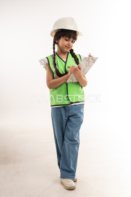 تدوين وكتابة الملاحظات والتخطيط للمشاريع، العمل في القطاع الهندسي، اختيار المستقبل منذ الصغر، بنت سعودية تكتب بالقلم بايماءات التركيز، بورتريه لفتاة عربية خليجية عمانية ترتدي الستره والخوذة تحمل بيدها اوراق خاصة بالمشروع، الطول الكامل للجسم، خلفية بيضاء