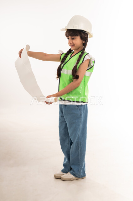 مفهوم العمل في القطاع الهندسي، تدقيق ومراجعة المخططات الهندسية، تحديد واختيار المستقبل منذ الصغر، بنت سعودية بايماءات الشغف لتحقيق حلم الطفولة، بورتريه لفتاة عربية خليجية عمانية ترتدي الستره وخوذة الحماية وتحمل مخطط المشروع، الطول الكامل للجسم، خلفية بيضاء