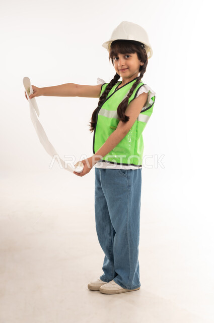 تدقيق ومراجعة المخططات الهندسية، تحديد واختيار المستقبل منذ الصغر، مفهوم العمل في القطاع الهندسي، بنت سعودية بايماءات الشغف لتحقيق حلم الطفولة، بورتريه لفتاة عربية خليجية عمانية ترتدي الستره وخوذة الحماية وتحمل مخطط المشروع، الطول الكامل للجسم، خلفية بيضاء