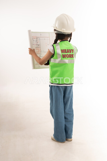مراجعة وتدقيق المخططات الهندسية، تحديد واختيار المستقبل منذ الصغر، مفهوم العمل في القطاع الهندسي، بنت سعودية بايماءات الشغف لتحقيق حلم الطفولة، بورتريه من الخلف لفتاة عربية خليجية عمانية ترتدي الستره وخوذة الحماية وتحمل مخطط المشروع، الطول الكامل للجسم، خلفية بيضاء