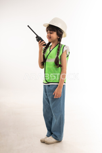 تحديد واختيار المستقبل منذ الصغر، مفهوم العمل في القطاع الهندسي، بنت سعودية بايماءات الشغف لتحقيق حلم الطفولة، بورتريه لفتاة عربية خليجية عمانية ترتدي الستره وخوذة الحماية تحمل جهاز الإتصال اللاسلكي وتقوم بالتحدث عبره، الطول الكامل للجسم، خلفية بيضاء