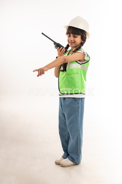 اختيار وتحديد المستقبل منذ الصغر، مفهوم العمل في القطاع الهندسي، بنت سعودية بايماءات الشغف لتحقيق حلم الطفولة، بورتريه لفتاة عربية خليجية عمانية ترتدي الستره وخوذة الحماية تحمل جهاز الإتصال اللاسلكي وتقوم بالإشارة إلى شيء ما، الطول الكامل للجسم، خلفية بيضاء