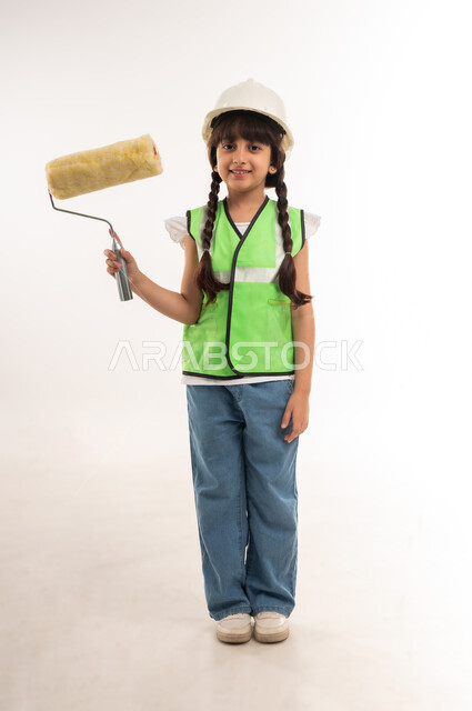 مفهوم العمل في القطاع الهندسي، أعمال الترميم وتشطيبات البناء، اختيار المستقبل منذ الصغر، بنت سعودية بايماءات الشغف لتحقيق حلم الطفولة، بورتريه لفتاة عربية خليجية عمانية ترتدي الستره وخوذة الحماية تحمل بيدها بكرة الدهان، الطول الكامل للجسم، خلفية بيضاء