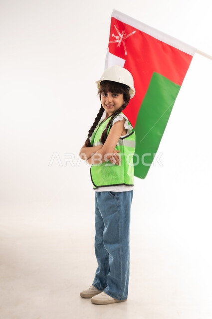 العمل في القطاع الهندسي، اختيار المستقبل منذ الصغر، ايماءات الشغف لتحقيق حلم الطفولة، بورتريه لفتاة عربية خليجية عمانية ترتدي الستره وخوذة الحماية تقف بايدي متقاطعة وخلفها العلم العماني، الفخر والإعتزاز بالهوية الوطنية، الطول الكامل للجسم، خلفية بيضاء