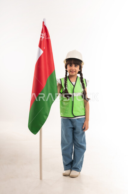 اختيار المستقبل منذ الصغر، مفهوم العمل في القطاع الهندسي، ايماءات الشغف لتحقيق حلم الطفولة، بورتريه لفتاة عربية خليجية عمانية ترتدي الستره وخوذة الحماية تقف باستقامة وتحمل العلم العماني، الفخر والإعتزاز بالهوية الوطنية، الطول الكامل للجسم، خلفية بيضاء