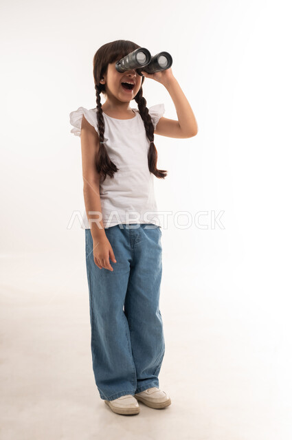 متعة الاستكشاف والمغامرة، رؤية الاشياء البعيدة، أهداف وطموحات مستقبلية، بورتريه لفتاة عربية خليجية عمانية تمسك بيدها منظار الرؤية ثنائي العينين لمشاهدة شيئا ما بايماءات السعادة والسرور، بنت سعودية تنظر عبر التلسكوب، صورة الجسم بالطول الكامل، خلفية بيضاء