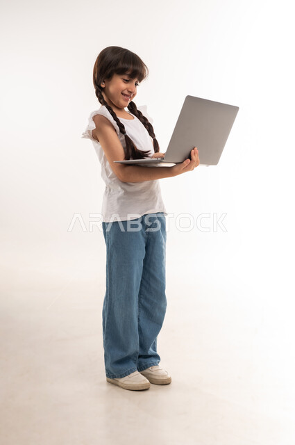 Browsing social media, children using modern technology, portrait of an Arab Gulf Omani girl standing straight and holding a laptop, Saudi girl making a video call with friends, e-learning in the Sultanate of Oman, full body length, white background