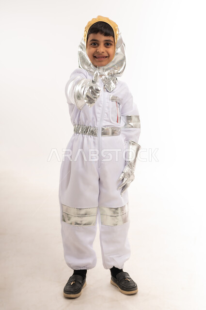 دعم قطاع الفضاء في سلطنة عمان، حلم الوصول إلى القمر، الجد والشغف لتحقيق الأهداف في المستقبل، بورتريه لفتى عربي خليجي عماني مبتسم يرتدي زي رائد الفضاء، صبي سعودي يقوم بتوجيه اصبع السبابة إلى الأمام بايماءات اللإشارة الى شخص ما، الطول الكامل للجسم، خلفية بيضاء