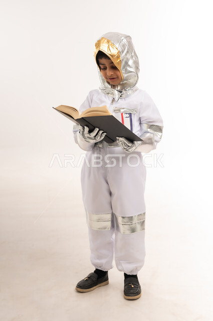التثقف والتعلم الذاتي، شغف التعرف على الكتب المختلفة، دعم قطاع الفضاء في سلطنة عمان، حلم الوصول إلى القمر، الجد في تحقيق أهداف المستقبل، بورتريه لفتى عربي خليجي عماني يرتدي زي رائد الفضاء، صبي سعودي يحمل بيده كتاب ويقوم بالقراءة، الطول الكامل للجسم، خلفية بيضاء