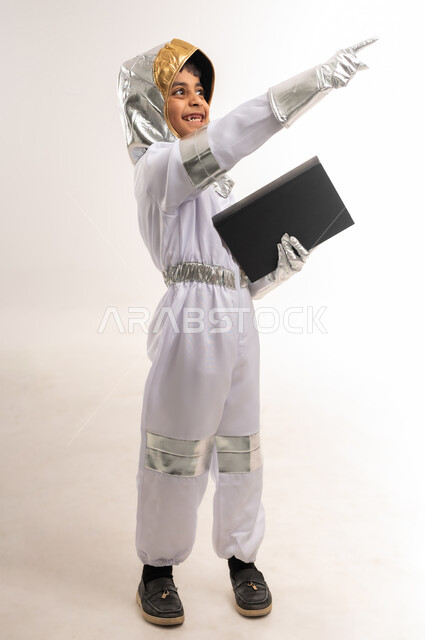 التعلم والتثقف الذاتي، شغف التعرف على الكتب المختلفة، دعم قطاع الفضاء في سلطنة عمان، حلم الوصول إلى القمر، الجد في تحقيق أهداف المستقبل، بورتريه لفتى عربي خليجي عماني يرتدي زي رائد الفضاء، صبي سعودي يحمل بيده كتاب ويشير الى شيء ما، الطول الكامل للجسم، خلفية بيضاء