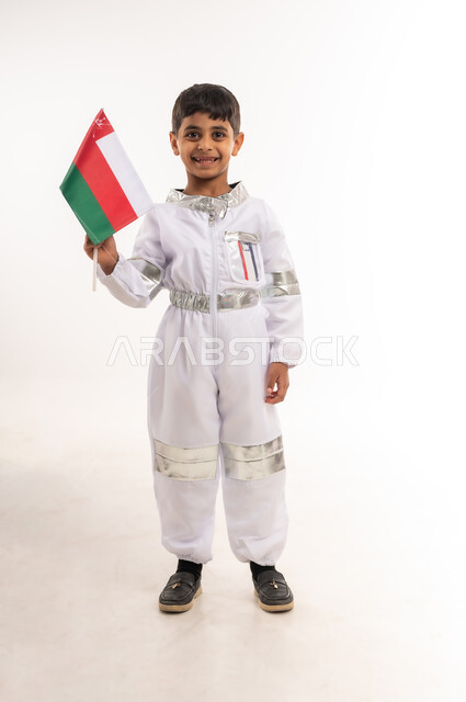 الشغف و الجد لتحقيق الأهداف في المستقبل، التطور و دعم قطاع الفضاء في سلطنة عمان، بورتريه لفتى عربي خليجي عماني مبتسم يرتدي زي رائد الفضاء ويحمل بيده العلم العماني، حلم الوصول إلى القمر، الفخر والإعتزاز بالهوية الوطنية، الطول الكامل للجسم، خلفية بيضاء