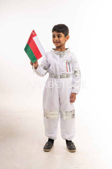 التطور و دعم قطاع الفضاء في سلطنة عمان، بورتريه لفتى عربي خليجي عماني مبتسم يرتدي زي رائد الفضاء ويحمل بيده العلم العماني، الشغف و الجد لتحقيق الأهداف في المستقبل، حلم الوصول إلى القمر، الفخر والإعتزاز بالهوية الوطنية، الطول الكامل للجسم، خلفية بيضاء