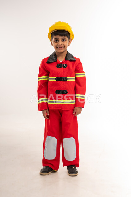 النظر إلى الكاميرا بايماءات السعادة والسرور، العمل في مجال الإنقاذ والحماية المدنية، التخطيط للمهن والوظائف المستقبلية، بورتريه لولد عربي خليجي عماني يرتدي الخوذة وسترة الحماية الخاصة برجال الإطفاء، صبي سعودي يقف باستقامة، الطول الكامل للجسم، خلفية بيضاء