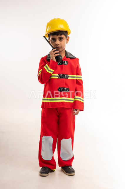 مفهوم العمل في مجال الانقاذ والحمايه المدنية، تحديد مهنة المستقبل، آمال وأمنيات لمهن و وظائف مستقبلية، بورتريه لولد عربي خليجي عماني يرتدي الخوذة وسترة الحماية الخاصة برجال الإطفاء، صبي سعودي يحمل بيده جهاز الإتصال اللاسلكي للاعلام عن حالة طارئة, الطول الكامل للجسم، خلفية بيضاء