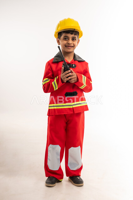تحديد مهنة المستقبل، مفهوم العمل في مجال الانقاذ والحمايه المدنية، آمال وأمنيات لمهن و وظائف مستقبلية، بورتريه لولد عربي خليجي عماني يرتدي الخوذة وسترة الحماية الخاصة برجال الإطفاء، صبي سعودي يحمل بيده جهاز الإتصال اللاسلكي للاعلام عن حالة طارئة, الطول الكامل للجسم، خلفية بيضاء