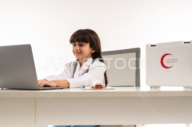 الأهداف والطموحات المستقبلية بالعمل في العيادات والمشافي، استعمال الاجهزة التقنية في المجال الطبي، اختيار وتحديد مهنة المستقبل منذ الصغر، بورتريه لبنت عربية خليجية عمانية ترتدي المعطف الأبيض، فتاة سعودية تجلس امام الطاولة تستخدم الحاسوب المحمول، خلفية بيضاء