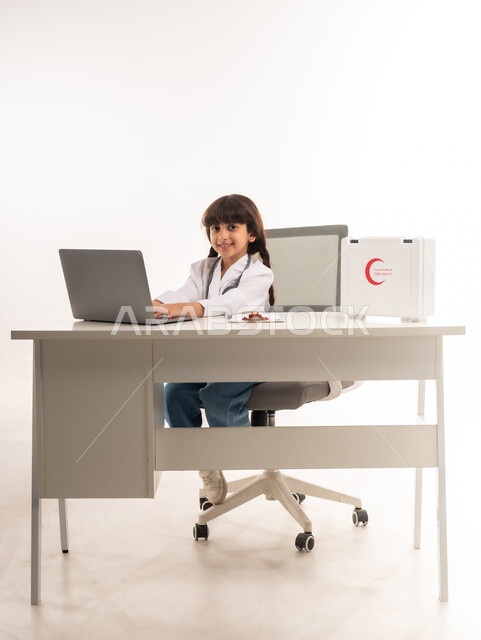 استخدام الاجهزة التقنية في المجال الطبي، اختيار وتحديد مهنة المستقبل منذ الصغر، الأهداف والطموحات المستقبلية بالعمل في العيادات والمشافي، بورتريه لبنت عربية خليجية عمانية ترتدي المعطف الأبيض، فتاة سعودية تجلس امام الطاولة تستخدم الحاسوب المحمول، خلفية بيضاء