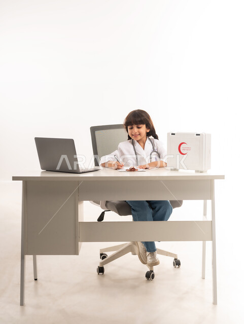 اختيار مهنة المستقبل، الأهداف والطموحات المستقبلية بالعمل في العيادات والمشافي، تدوين نتائج الفحوصات بالسجل الطبي، بورتريه لبنت عربية خليجية عمانية ترتدي المعطف الأبيض، فتاة سعودية تجلس امام الطاولة تقوم بإعداد التقارير الطبيه وبجانبها الحاسوب المحمول، خلفية بيضاء