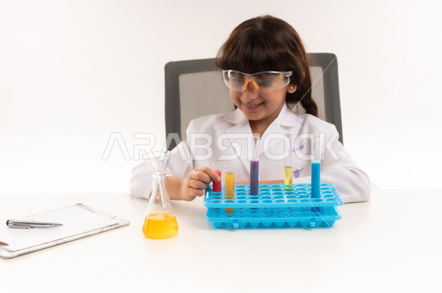Choosing and determining the future profession since childhood, the wish to obtain the profession of a chemical researcher, conducting experiments and biochemical reactions in the laboratory, a portrait of an Arab Gulf Omani girl wearing a white coat and using test tubes, a Saudi girl conducting scientific research in the chemistry laboratory, white background