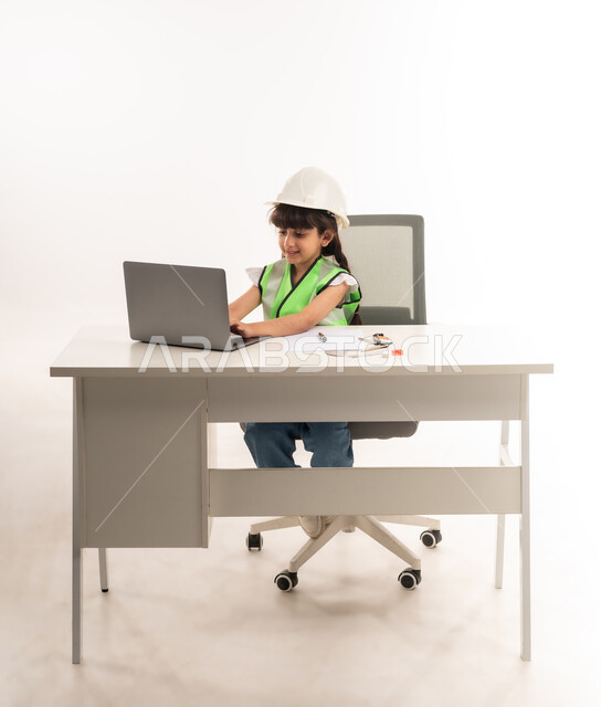 Saudi girl with gestures of passion to achieve her childhood dream, working in the engineering sector in the Sultanate of Oman, choosing the future from a young age, using modern technical devices in the engineering sector, portrait of an Arab Gulf Omani girl wearing a jacket sitting in front of a table using a laptop, white veil