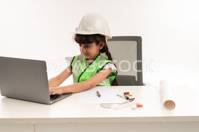Saudi girl with gestures of passion to achieve her childhood dream, working in the engineering sector in the Sultanate of Oman, choosing the future from a young age, using modern technical devices in the engineering sector, portrait of an Arab Gulf Omani girl wearing a jacket sitting in front of a table using a laptop, white veil
