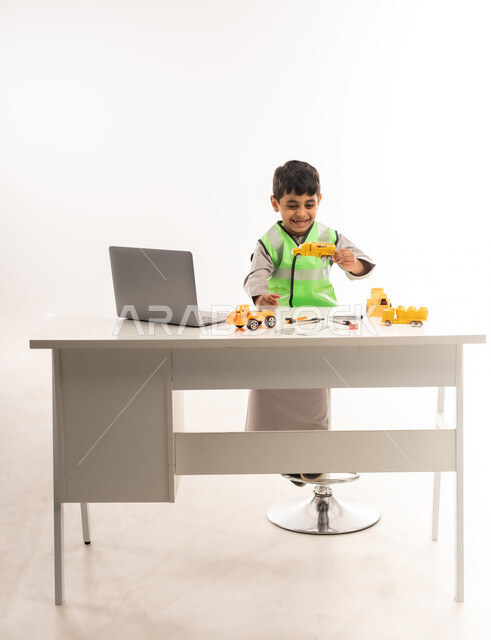 Gestures of passion to achieve a childhood dream, using modern technical devices in the engineering sector, portrait of an Arab Gulf Omani boy wearing a dishdasha and a jacket sitting in front of a table using a laptop, working in the engineering sector in the Sultanate of Oman, choosing and determining the future from a young age, white cloak