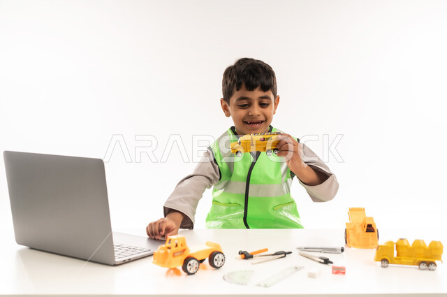 Gestures of passion to achieve a childhood dream, using modern technical devices in the engineering sector, portrait of an Arab Gulf Omani boy wearing a dishdasha and a jacket sitting in front of a table using a laptop, working in the engineering sector in the Sultanate of Oman, choosing and determining the future from a young age, white cloak