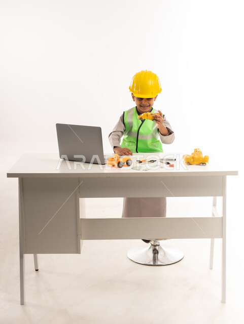 Working in the engineering sector in the Sultanate of Oman, using modern technical devices in the engineering sector, gestures of passion to achieve a childhood dream, portrait of an Arab Gulf Omani boy wearing a dishdasha and a jacket sitting in front of a table using a laptop, choosing and determining the future from a young age, white cloak