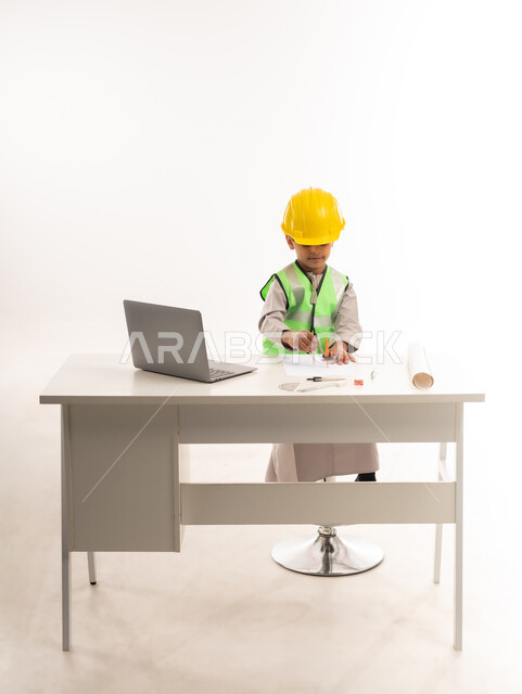 Using modern technical devices in the engineering sector, gestures of passion to achieve a childhood dream, portrait of an Arab Gulf Omani boy wearing a dishdasha and a jacket sitting in front of a table using a laptop, working in the engineering sector in the Sultanate of Oman, choosing and determining the future from a young age, white cloak