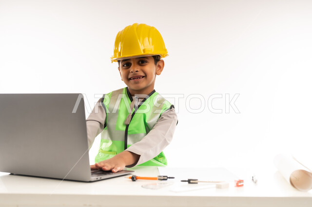 Working in the engineering sector in the Sultanate of Oman, using modern technical devices in the engineering sector, gestures of passion to achieve a childhood dream, portrait of an Arab Gulf Omani boy wearing a dishdasha and a jacket sitting in front of a table using a laptop, choosing and determining the future from a young age, white cloak