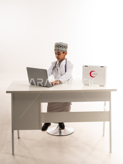 الطموحات والأهداف المستقبلية في العمل في العيادات والمشافي، استخدام الاجهزة التقنية الحديثة في المجال الطبي، بورتريه لولد عربي خليجي عماني يرتدي الكمة والمعطف الأبيض يجلس امام الطاولة ويستخدم الحاسوب المحمول، تحديد واختيار مهنة المستقبل منذ الصغر، خلفية بيضاء