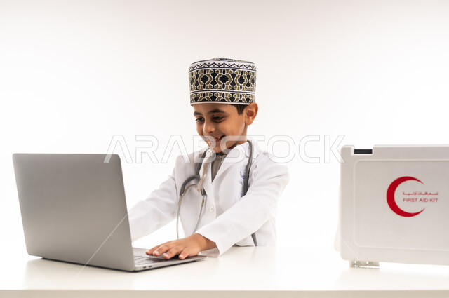 استخدام الاجهزة التقنية الحديثة في المجال الطبي، الطموحات والأهداف المستقبلية في العمل في العيادات والمشافي، بورتريه لولد عربي خليجي عماني يرتدي الكمة والمعطف الأبيض يجلس امام الطاولة ويستخدم الحاسوب المحمول، تحديد واختيار مهنة المستقبل منذ الصغر، خلفية بيضاء