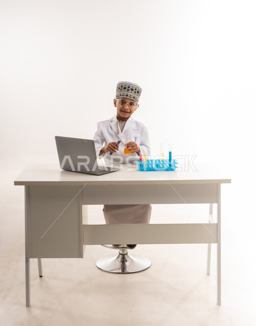 استخدام أنابيب الأختبار وتدوين النتائج على الحاسب المحمول، مهنة اخصائي مختبرات حيوية، اختيار وتحديد المستقبل منذ الصغر، بورتريه لولد عربي خليجي عماني يرتدي الكمة والمعطف الأبيض يجلس امام الطاولة يضع نظارات واقية لإجراء التفاعلات الكيميائية، خلفية بيضاء