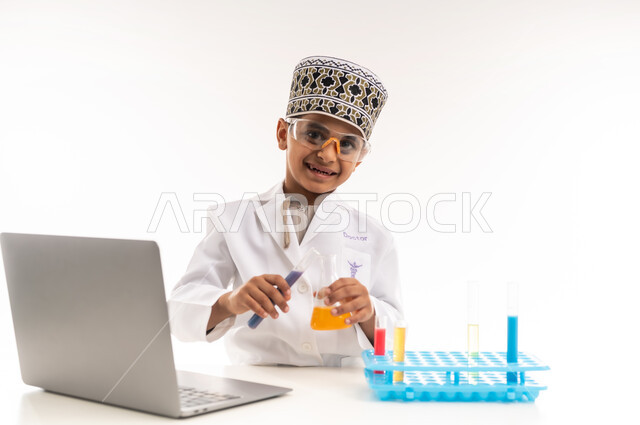 مهنة اخصائي مختبرات حيوية، اختيار وتحديد المستقبل منذ الصغر، بورتريه مقرب لولد عربي خليجي عماني يرتدي الكمة والمعطف الأبيض يجلس امام الطاولة يضع نظارات واقية لإجراء التفاعلات الكيميائية، استخدام أنابيب الأختبار وتدوين النتائج على الحاسب المحمول، خلفية بيضاء