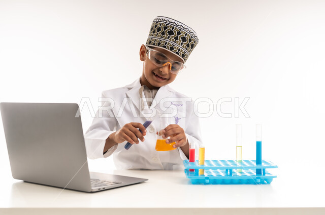 Choosing and determining the future from a young age, the profession of a bio-laboratory specialist, a close-up portrait of an Arab Gulf Omani boy wearing a turban and a white coat sitting in front of a table wearing protective glasses to conduct chemical reactions, using test tubes and writing down the results on a laptop, white background