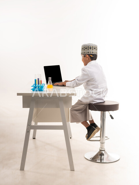 اختيار وتحديد المستقبل منذ الصغر، مهنة اخصائي مختبرات حيوية، بورتريه من الجانب لولد عربي خليجي عماني يرتدي الكمة والمعطف الأبيض يجلس امام الطاولة يضع نظارات واقية لإجراء التفاعلات الكيميائية، استخدام أنابيب الأختبار وتدوين النتائج على الحاسب المحمول، خلفية بيضاء