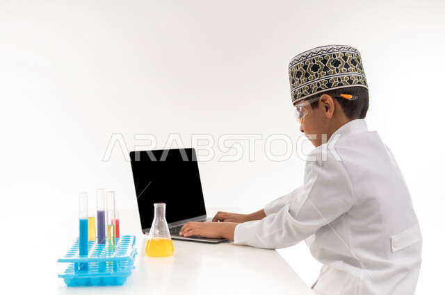 مهنة اخصائي مختبرات حيوية، اختيار وتحديد المستقبل منذ الصغر، بورتريه مقرب لولد عربي خليجي عماني يرتدي الكمة والمعطف الأبيض يجلس امام الطاولة يضع نظارات واقية لإجراء التفاعلات الكيميائية، استخدام أنابيب الأختبار وتدوين النتائج على الحاسب المحمول، خلفية بيضاء