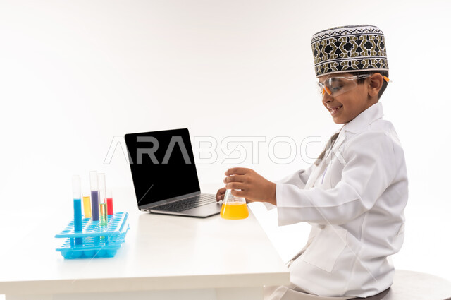 اختيار وتحديد المستقبل منذ الصغر، مهنة اخصائي مختبرات حيوية، بورتريه مقرب لولد عربي خليجي عماني يرتدي الكمة والمعطف الأبيض يجلس امام الطاولة يضع نظارات واقية لإجراء التفاعلات الكيميائية، استخدام أنابيب الأختبار وتدوين النتائج على الحاسب المحمول، خلفية بيضاء