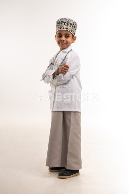 الوقوف بأيدي متقاطعة بايماءات الثقة بالنفس لتحقيق حلم الطفوله، تقديم الخدمات الصحية، بورتريه لولد عربي خليجي عماني يرتدي الكمة والمعطف الأبيض والسماعات الطبية يحلم بأن يصبح طبيب عام، اختيار وتحديد المهنة المستقبلية منذ الصغر، صورة الجسم بالطول الكامل، خلفية بيضاء
