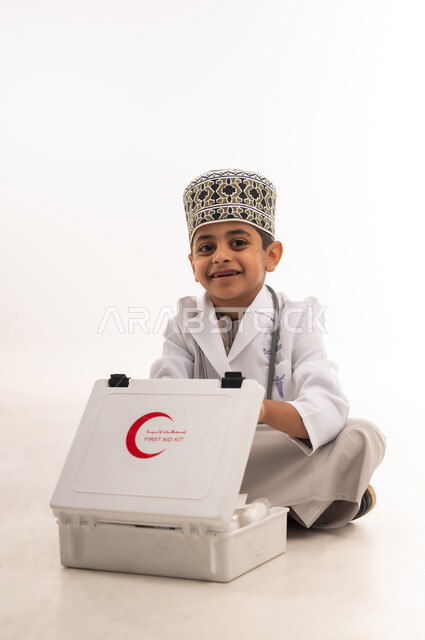 النظر الى الكاميرا بتعابير الشغف لتحقيق حلم التمريض، تقديم الرعاية والخدمات الصحية، بورتريه لفتى عربي خليجي عماني مبتسم يرتدي الكمة والمعطف الأبيض يجلس على الارض يتفقد محتويات حقيبة الاسعافات الأولية، اختيار وتحديد المهنة المستقبلية منذ الصغر، خلفية بيضاء