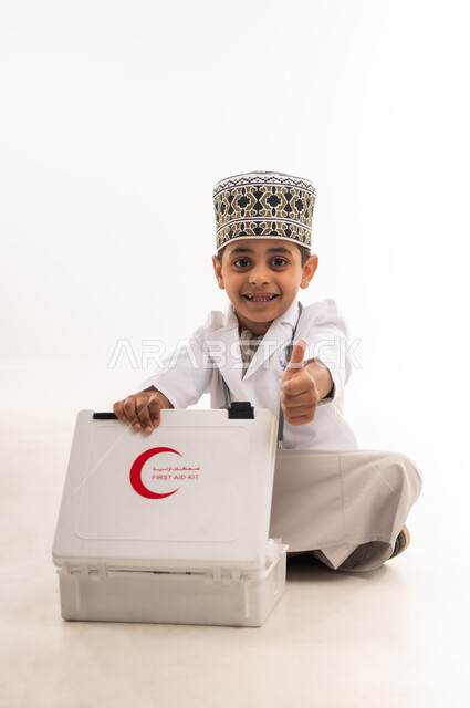 النظر الى الكاميرا بتعابير الشغف والاعجاب، تقديم الخدمات الصحية، بورتريه لفتى عربي خليجي عماني يرتدي الكمة والمعطف الأبيض يجلس على الارض امام حقيبة الاسعافات الأولية يرفع اصبع الابهام بايماءات القبول والرضا، اختيار وتحديد المهنة المستقبلية منذ الصغر، خلفية بيضاء