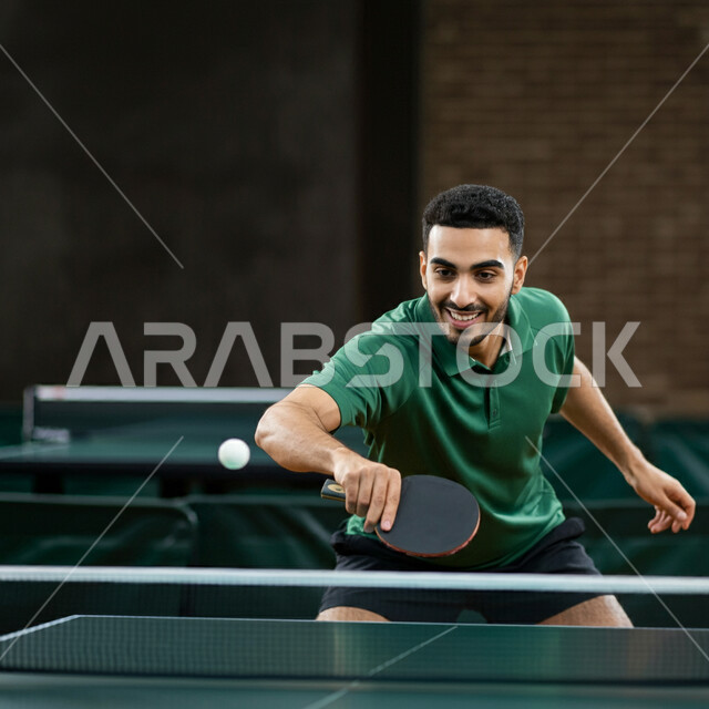 ممارسة رياضات وأنشطة بدنية ممتعة، شاب عربي خليجي سعودي يرتدي ملابس رياضية يحمل بيده مضرب تنس الطاولة، اللعب بسرعة وتركيز عالي، المحافظة على الرياضة والرشاقة الجسدية، الاستمتاع بالمباريات الودية والبطولات المحلية