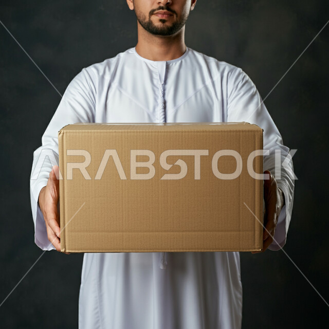 طلب المنتجات أونلاين, مفهوم التسوق الالكتروني، خدمات التوصيل السريع, الاعتماد على عمليات الشراء عبر الانترنت, بورتريه مقرب لرجل عربي خليجي إماراتي يرتدي الكندورة يحمل بيديه الصندوق, إيصال الطلبات إلى الزبائن بسهولة وسرعة، خلفية سوداء